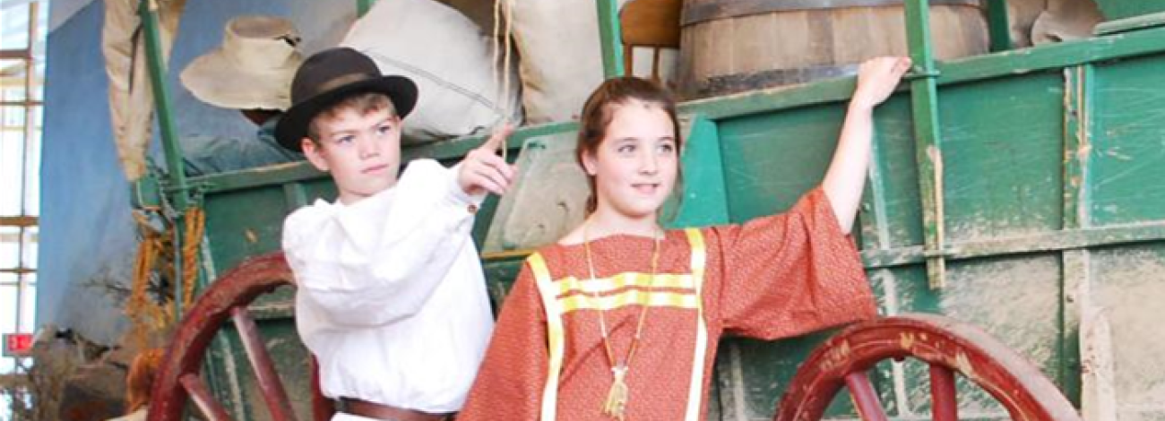 two children in old fashioned garb standing next to display of a covered wagon