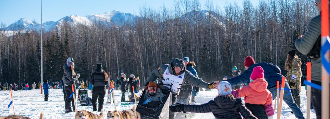 This picture depicts a team of sled dogs and a musher riding in the snow and greeting spectators underneath a blue sky.