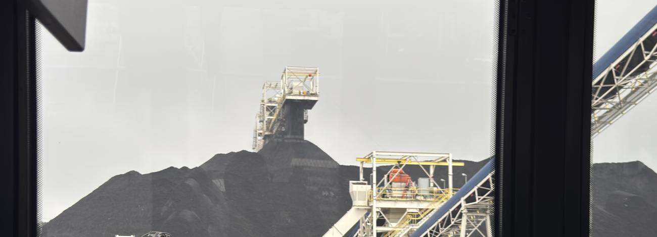 View from a vehicle showing heavy machinery and large coal piles at an industrial site, with conveyor belts and cloudy sky.