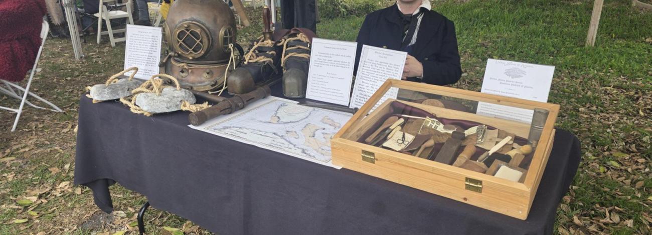 Outdoor display table with antique diving helmet, boots, weights, nautical tools, maps, and informational signs at a historical event.