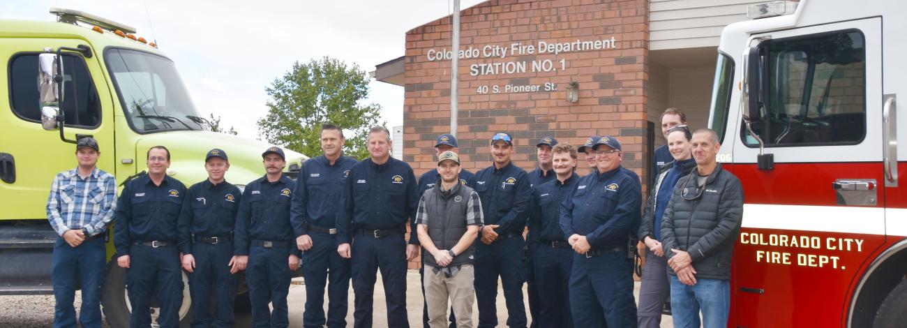 Firefighters in uniform stand in front of two large fire trucks one is green the other is red