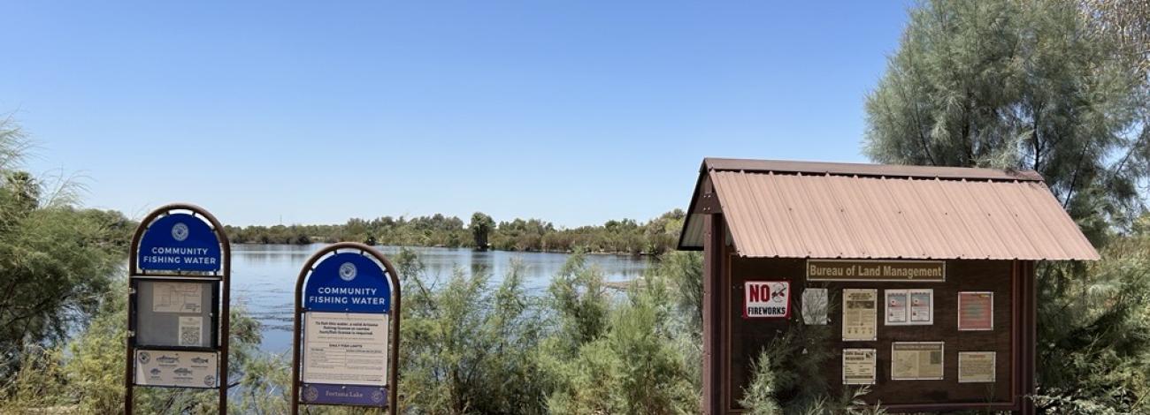 Two signs next to a calm pond with a brown wooden BLM kiosk to the right.