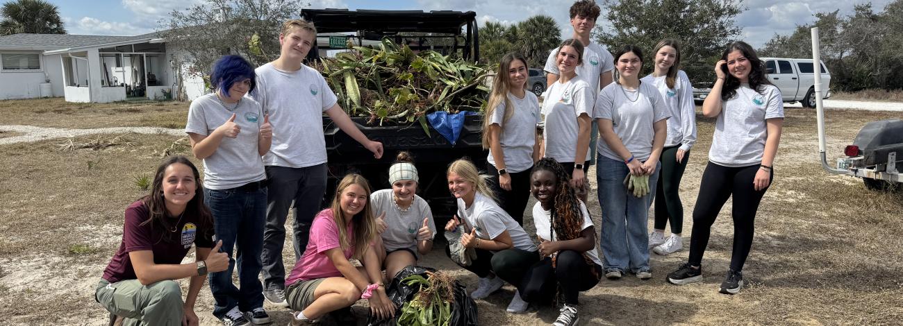 Group of young volunteers posing outdoors with collected plants and yard waste on a sunny day.