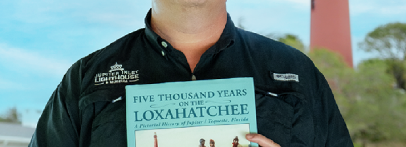 Person holding a book titled "Five Thousand Years on the Loxahatchee" with a lighthouse and trees in the background.