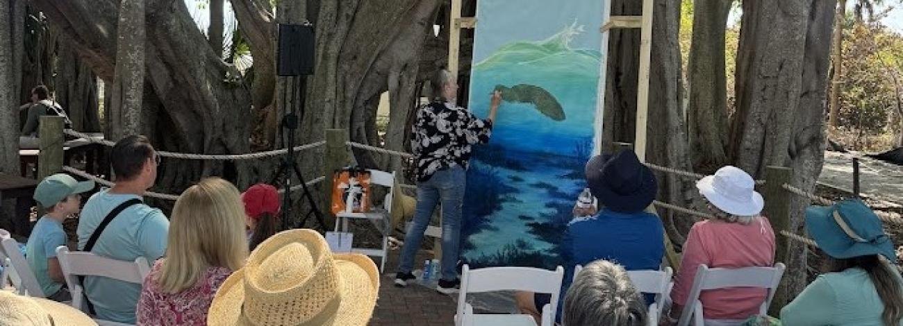 Artist painting a marine scene on a large canvas outdoors, with an audience seated on white chairs under large tree branches.