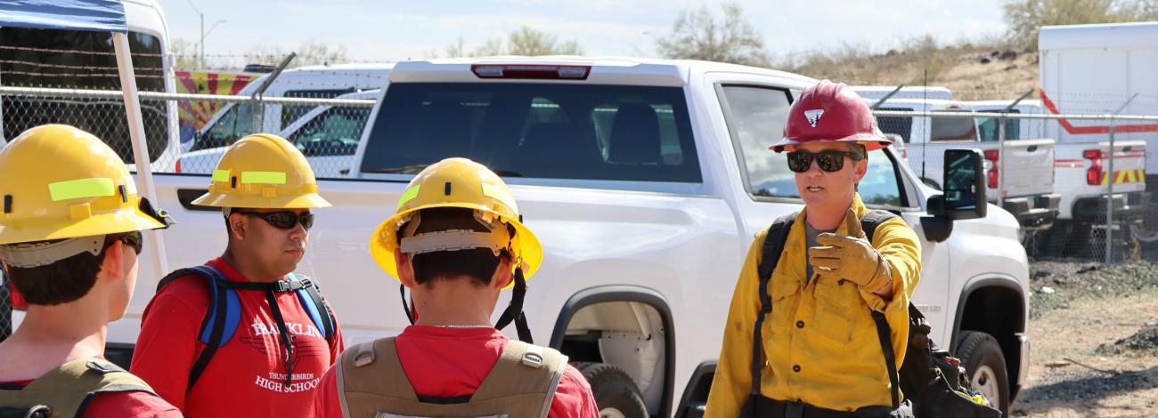 a wildland firefighter in yellow shirt, green pants, and hard hat, speaks to high school students