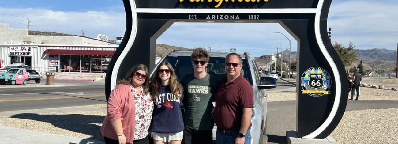 Four people stand in front of a large Route 66 Kingman roadside sign with a vintage vehicle display behind them on a sunny day.