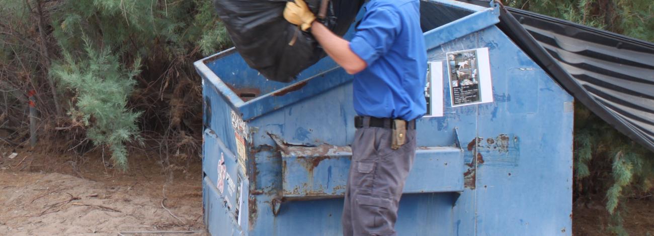 A man throws a bag of trash into a blue dumpster.