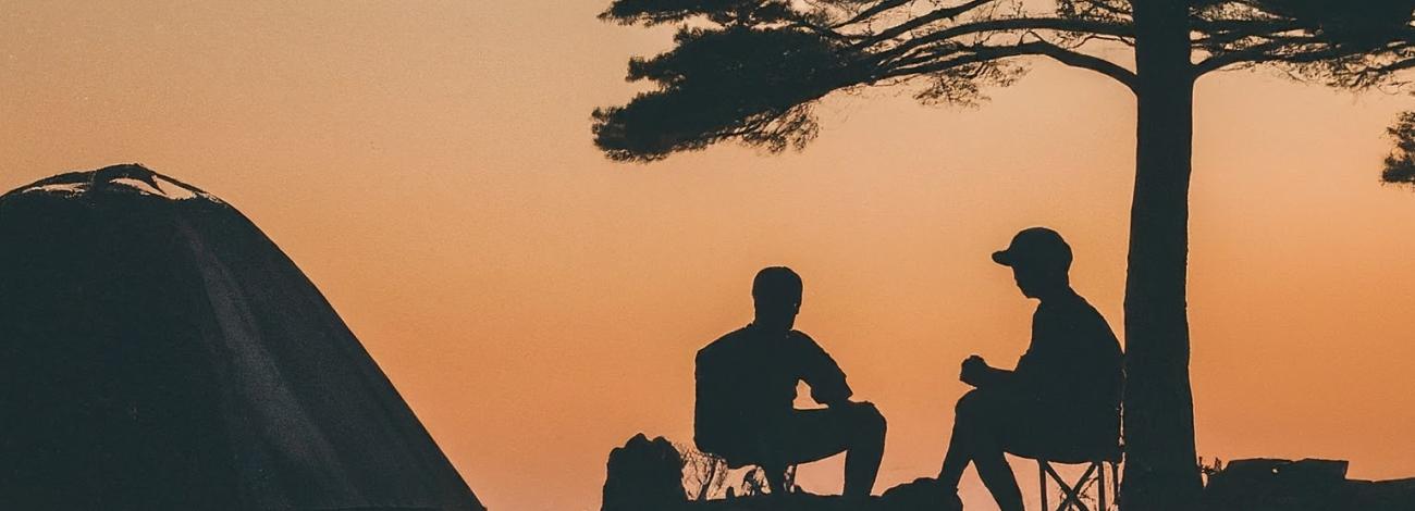 A photo of two people sitting under tree, near a tent