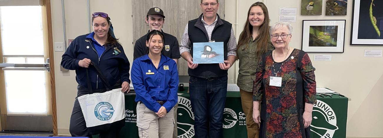 Group of six people standing indoors in front of tables with Federal Junior Duck Stamp banners, one person holding a duck painting.