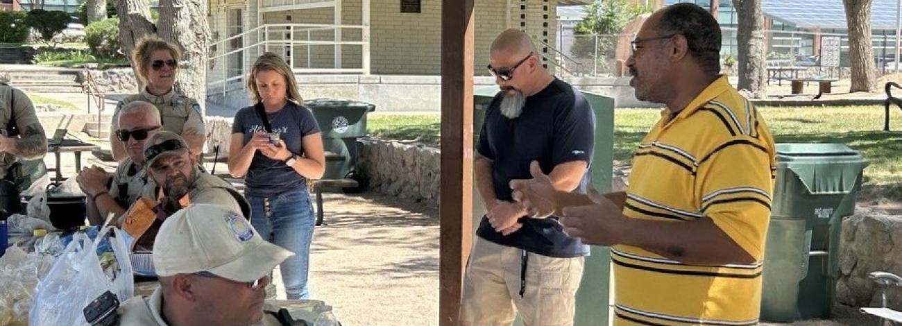 Several Bureau of Land Management staff members gather under a shaded pavilion while a man in a yellow striped shirt speaks to the group during an outdoor employee event.