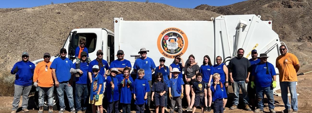 A Yuma County Public Works truck and a large roll-off dumpster sit in a desert cleanup area as volunteers collect trash across rocky public lands near Yuma, Arizona.