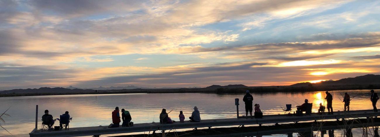 People sitting and standing on a dock at sunrise overlooking calm lake waters with colorful clouds reflecting on the surface.
