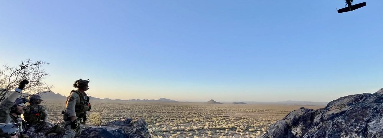 Tactical officers on a rocky ridge signal to an approaching helicopter during a desert rescue training exercise.