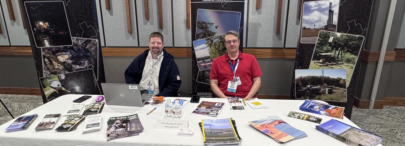 Two men seated behind a table with brochures at a U.S. Department of the Interior Bureau of Land Management informational booth.