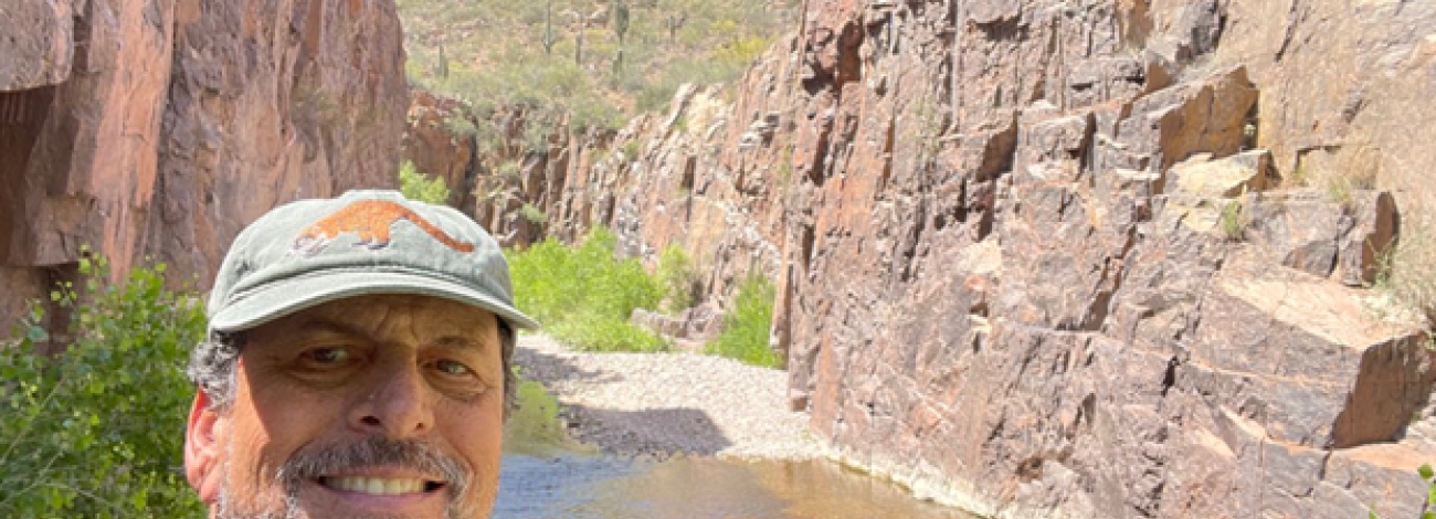 Hiker wearing a hat standing beside a narrow desert canyon stream, with tall rock walls rising on both sides under a bright blue sky.