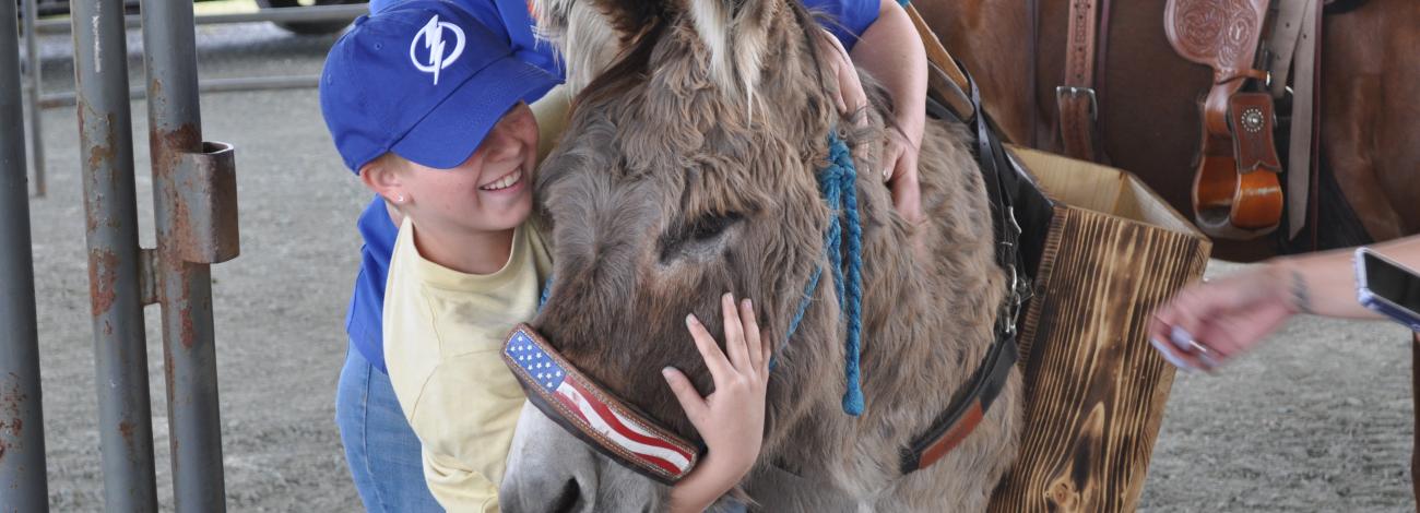 Child hugging a donkey wearing an American flag noseband, with a smiling adult behind and a saddled horse nearby.