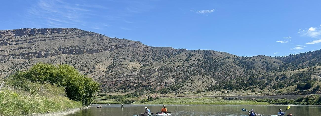 People in kayaks float on a river with hills in the background under a blue sky