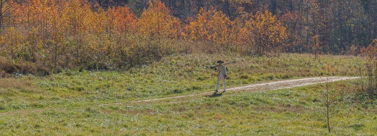 Person dressed in colonial attire walking along a path in a grassy field with autumn-colored trees in the background.