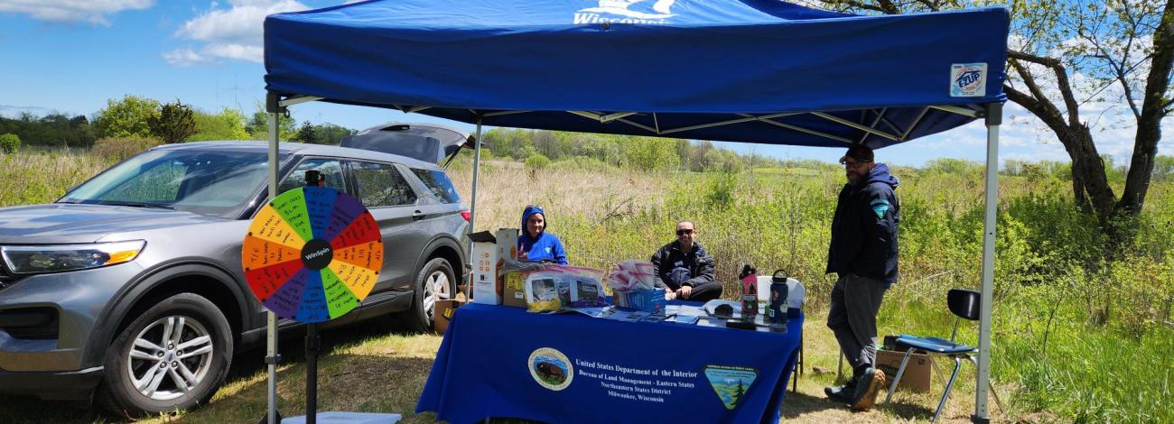 Outdoor event booth under a blue canopy with a colorful prize wheel and informational table near a silver SUV in a grassy field.