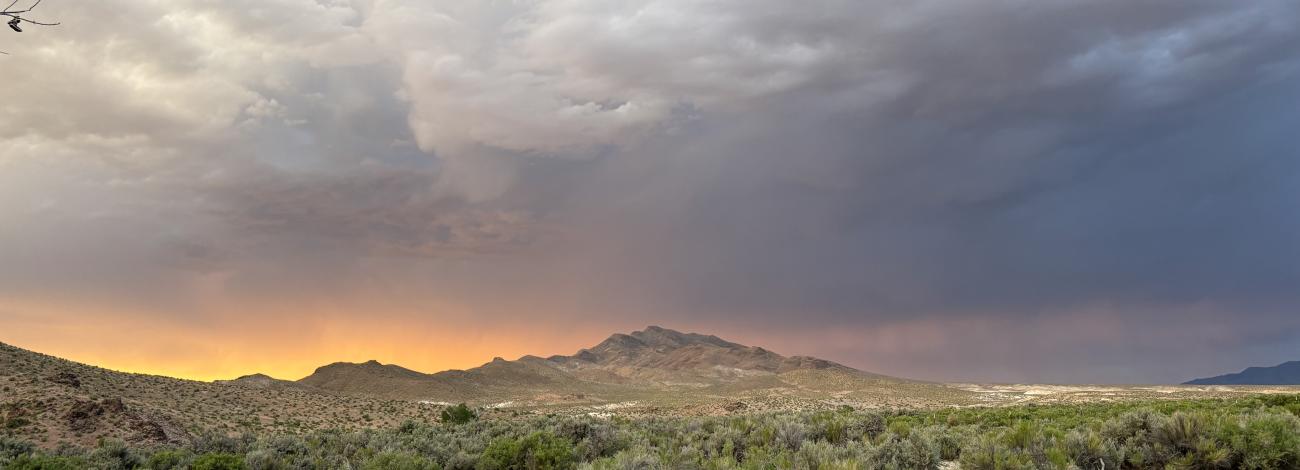 A dramatic desert landscape at sunset shows rugged hills and dense sagebrush in the foreground. The sky is filled with thick, layered storm clouds, shifting from deep gray to soft blue, while a bright band of orange light glows along the horizon, illuminating the mountains beneath it.