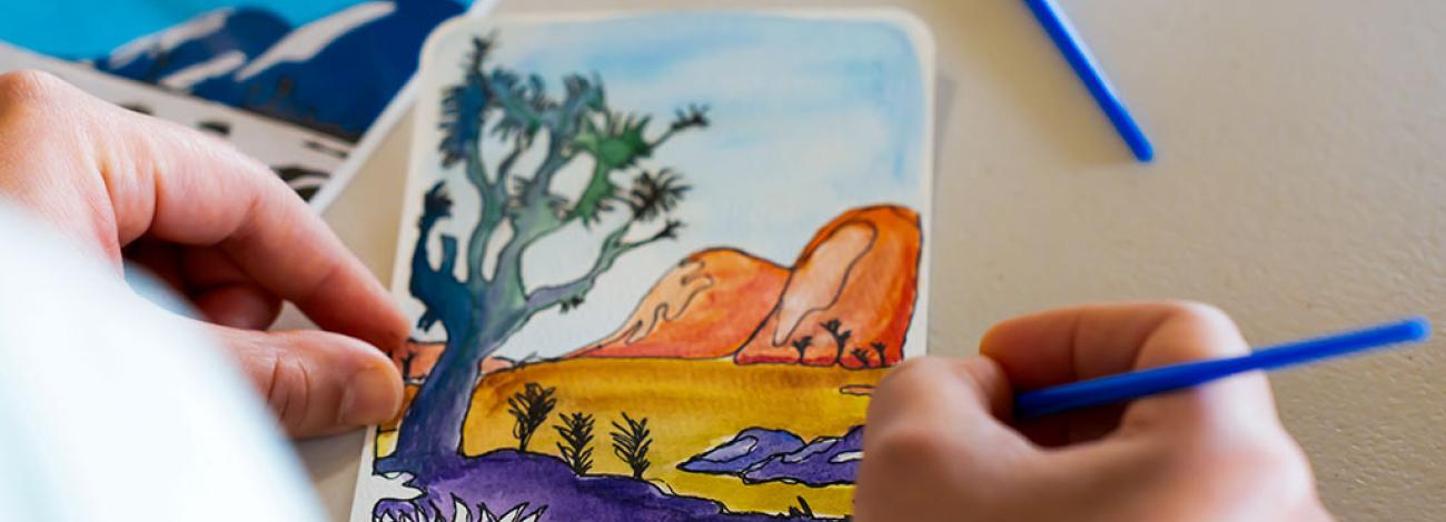 A person holds a small postcard-sized watercolor painting depicting a desert landscape with orange rock formations, a large tree in the foreground, and purple and yellow plants along the ground. The person uses a blue paintbrush to add details, with other painted cards and supplies visible on the table.