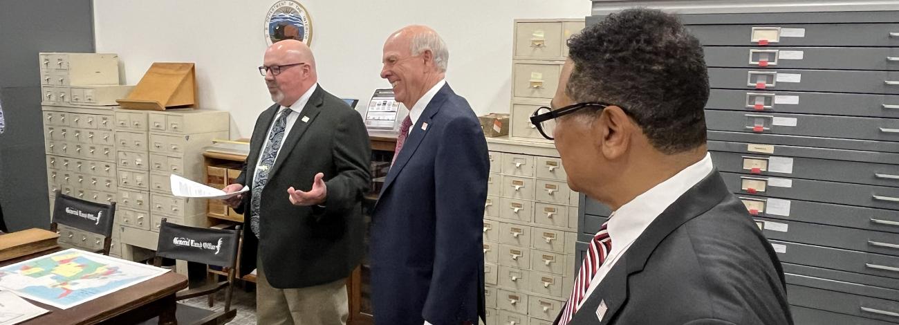Three people stand together in a room with map drawers and archival furniture. One person speaks while holding papers, and the others listen attentively. Colorful documents and historical materials are laid out on nearby tables.