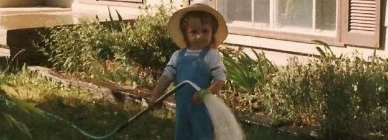 A small child standing in a yard, wearing overalls and a wide-brimmed hat, holding a garden hose that sprays water across the grass near a house.