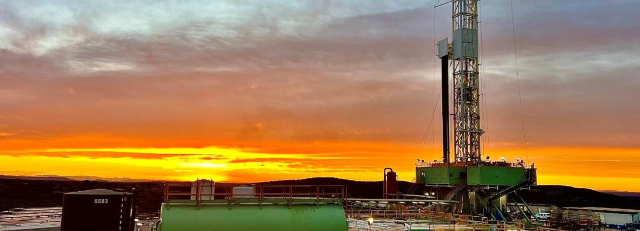 An onshore oil and gas drilling rig stands against a vibrant orange and blue sunrise sky. In the foreground, large green industrial mud tanks and a vertical black storage tank sit on a dirt lot. The tall metal derrick tower rises on the right side of the frame with its working lights illuminated.