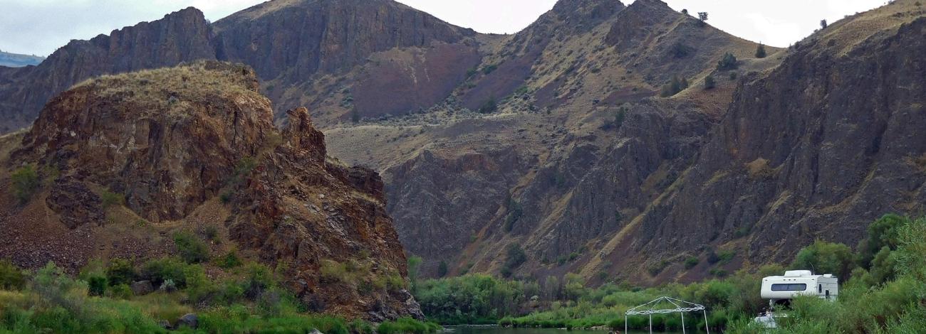 Riverbank campsite with two people sitting under a canopy beside a parked RV, set against steep rocky hills and a calm river.