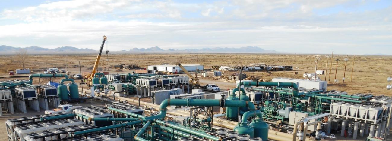 The image shows a large geothermal plant in an open field of dead grass under a cloudy sky.