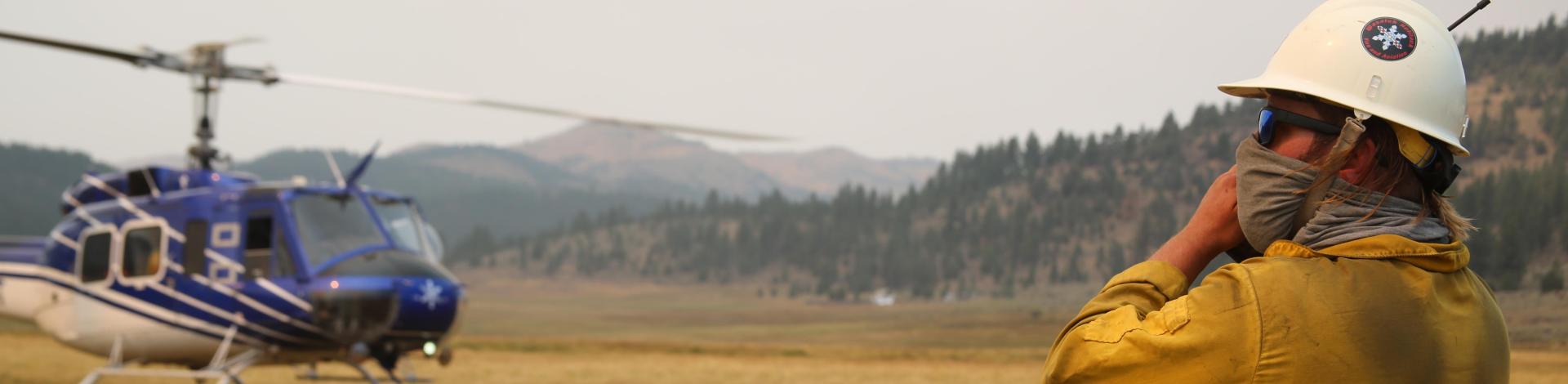 A man in a field talks into a handheld radio with a helicopter in the background