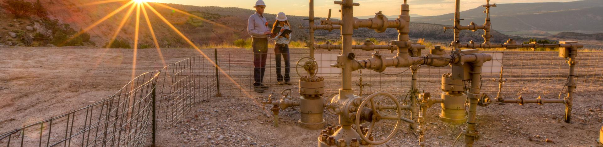 Two BLM employees work near oil and gas pipes with a mountain and the setting sun in the background.