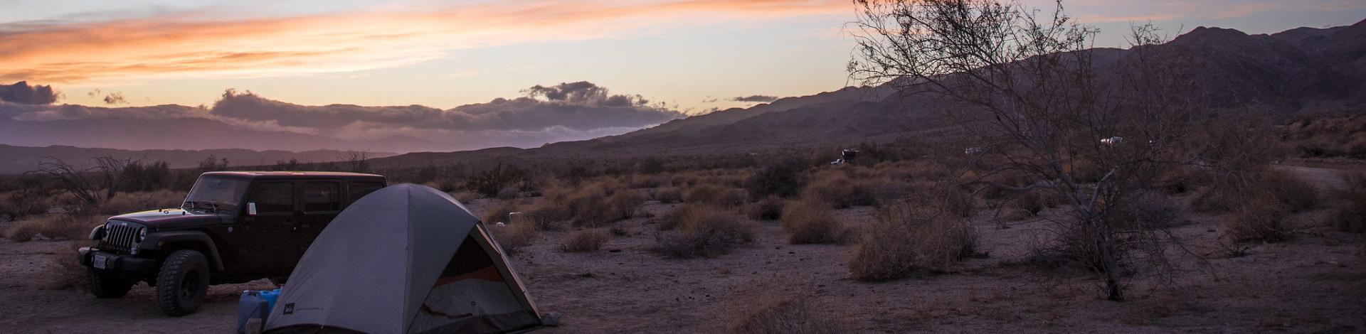 A jeep is parked next to a tent on a desert landscape, with a mountain in the background and the setting sun turning the sky and clouds pink and blue.