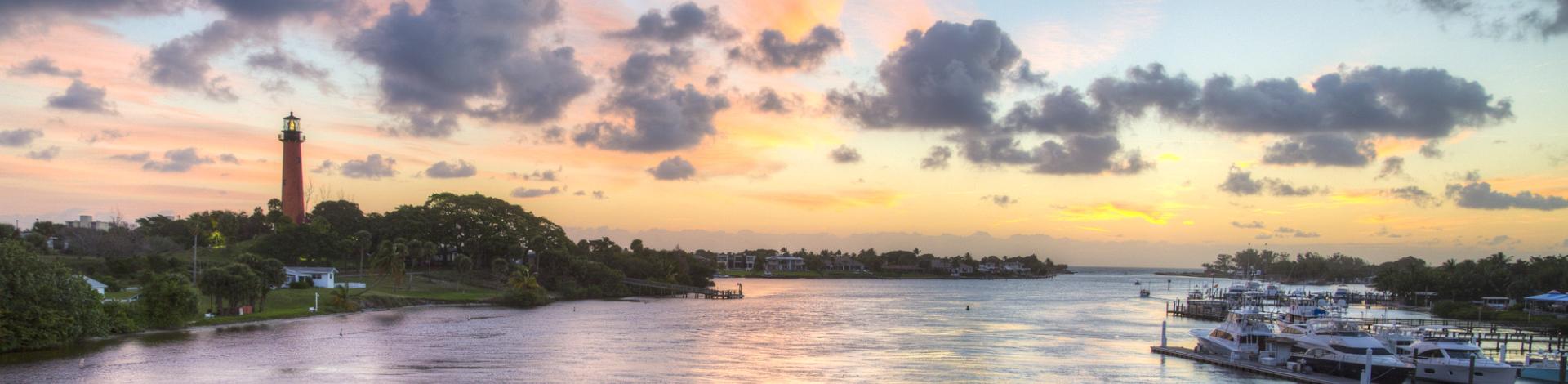 Sunset over a calm river with a lighthouse on the left and boats docked on the right under a colorful sky.