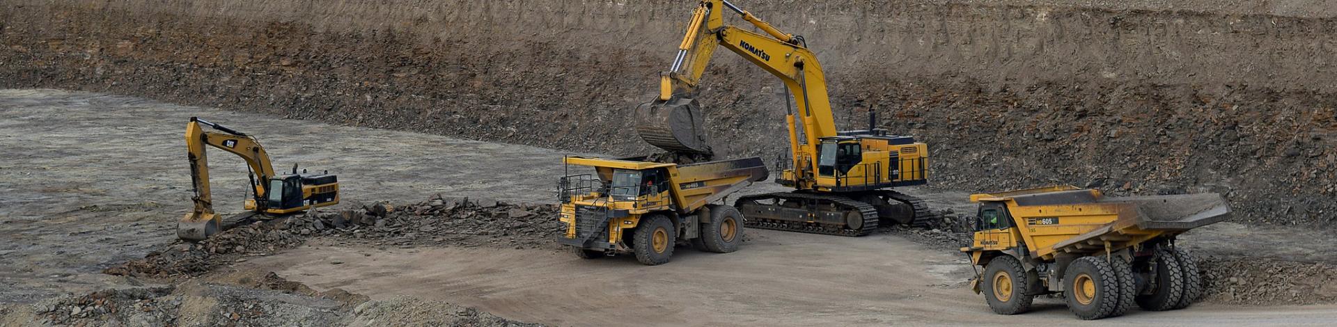 An excavator loads material into a large dump truck as another dump truck waits to be loaded.