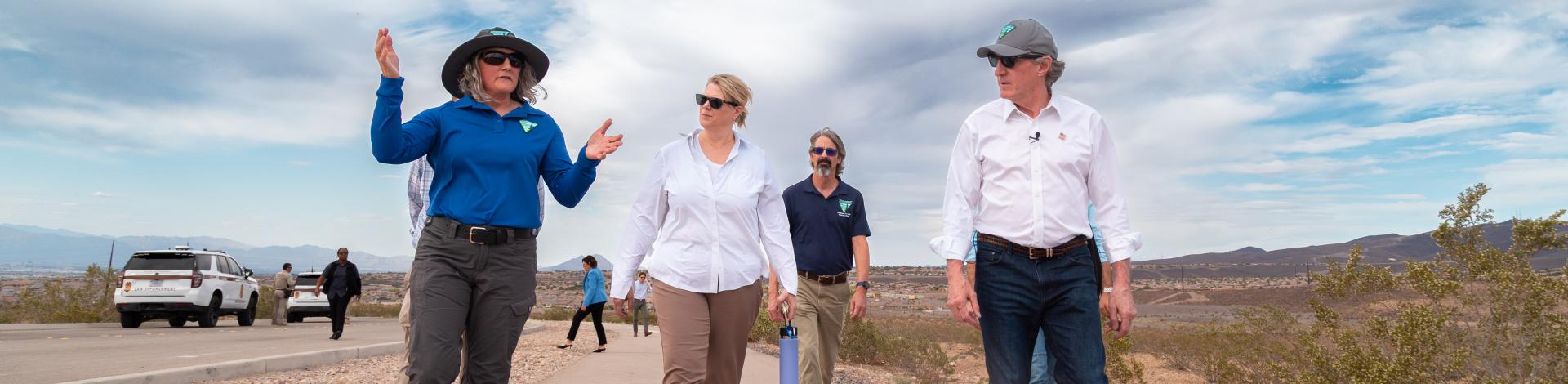 A woman in a blue BLM shirt gestures as she walks on a dirt road with Interior Secretary Doug Burgum and two other people, with a cloudy sky and official vehicles in the background.