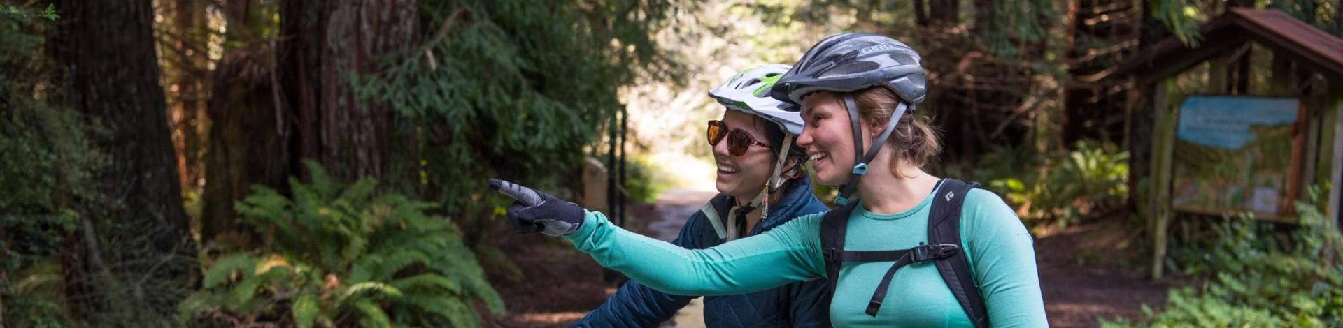 Two women with mountain bikes stoping at an interpretive panel on a forested trail.
