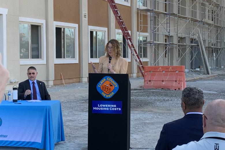 A woman in a tan suit stands behind a black podium in front of a brown apartment building that is under construction. A man in a suit sits at a table off to one side of the podium. The backs of the heads and shoulders of two other men seated in the audience are visible in the foreground.