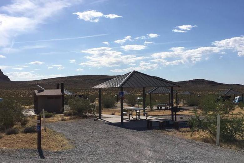 A photo showing a gravel parking spot next to a covered picnic table. A concrete path leads to a double-vault toilet, several other campsites with covered picnic tables can be seen in the background.  