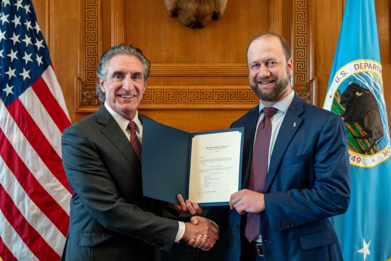 Two men in suits shake hands while holding a certificate in a formal wood-paneled room with U.S. flags behind them.