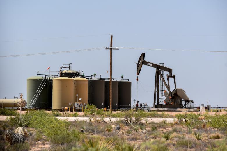 Oil pumpjack operating next to large storage tanks in a dry, desert landscape under a clear sky.