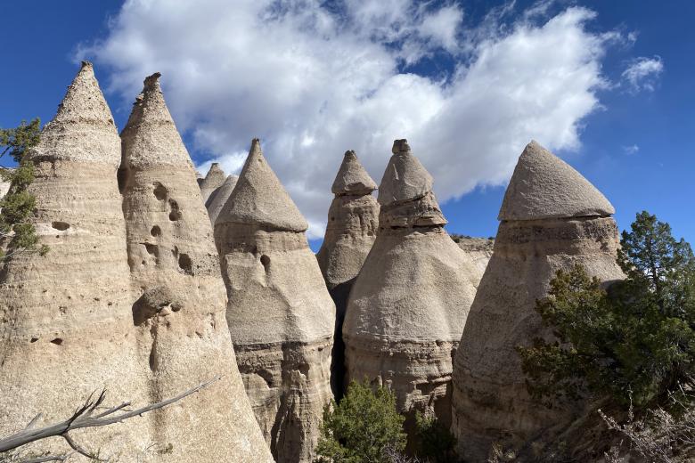 Cone-shaped rock formations, known as hoodoos, rise beneath a blue sky with scattered clouds and surrounding sparse vegetation.