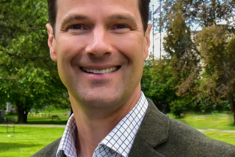 Kevin Pendergast, wearing a blue shirt and brown jacket, smiles in front of a green field.