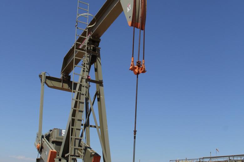 Large oil pumpjack operating under a clear blue sky in a desert-like industrial site with storage tanks in the background.