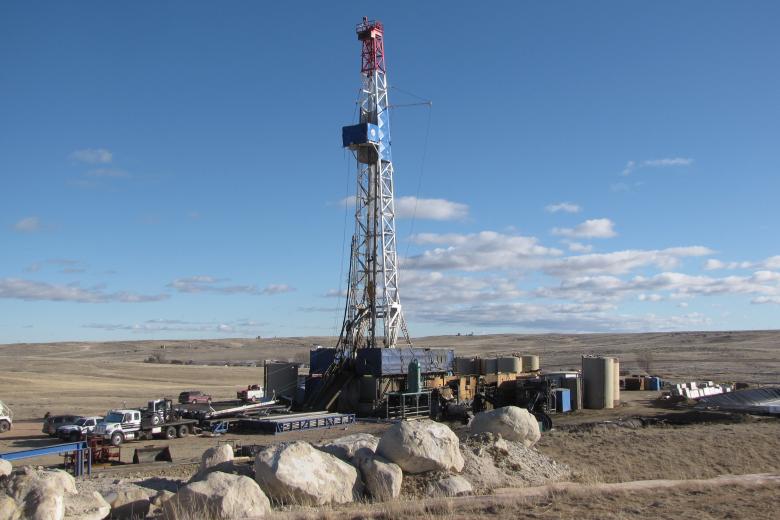 Oil drilling rig set against a clear blue sky with scattered clouds. The foreground features large rocks and vehicles.