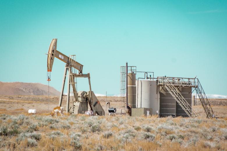 An oil pumpjack and storage tanks sit in a dry desert landscape with sparse grasses. A pronghorn antelope grazes near the base of the pumpjack under a bright blue sky.
