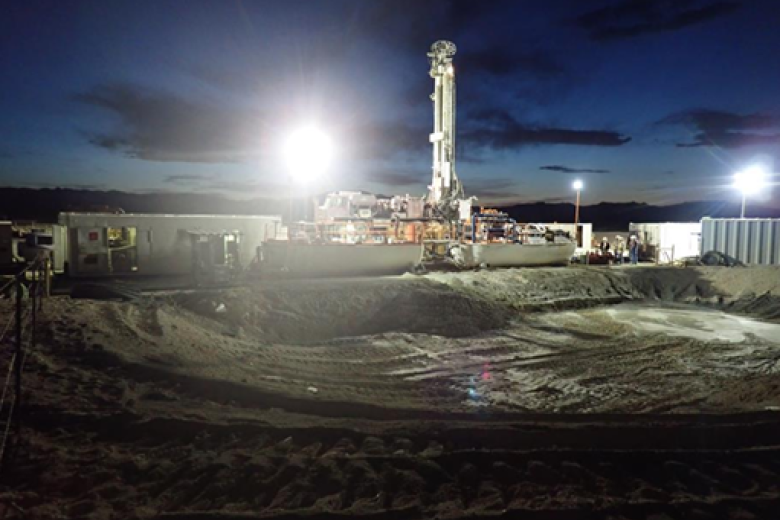 A white oil drill stands above several office trailers above an excavated pit about 50 meters wide and 5 meters deep. It is night. The scene is lit by 3 industrial light towers.