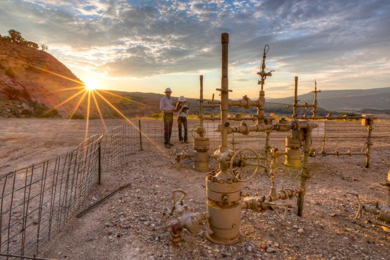 Two BLM employees work near oil and gas pipes with a mountain and the setting sun in the background.