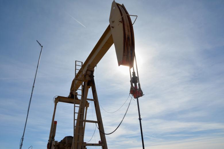 A low-angle shot captures the dark silhouette of a large oil pumpjack against a bright, sunlit sky. The sun is directly behind the pumpjack's horsehead, creating a brilliant flare and obscuring its details, while thin, wispy clouds streak across the blue sky. A thin antenna tower stands to the left of the machinery.